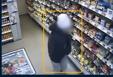 A store aisle camera frame with a detection box drawn around a shopper standing at the shelves