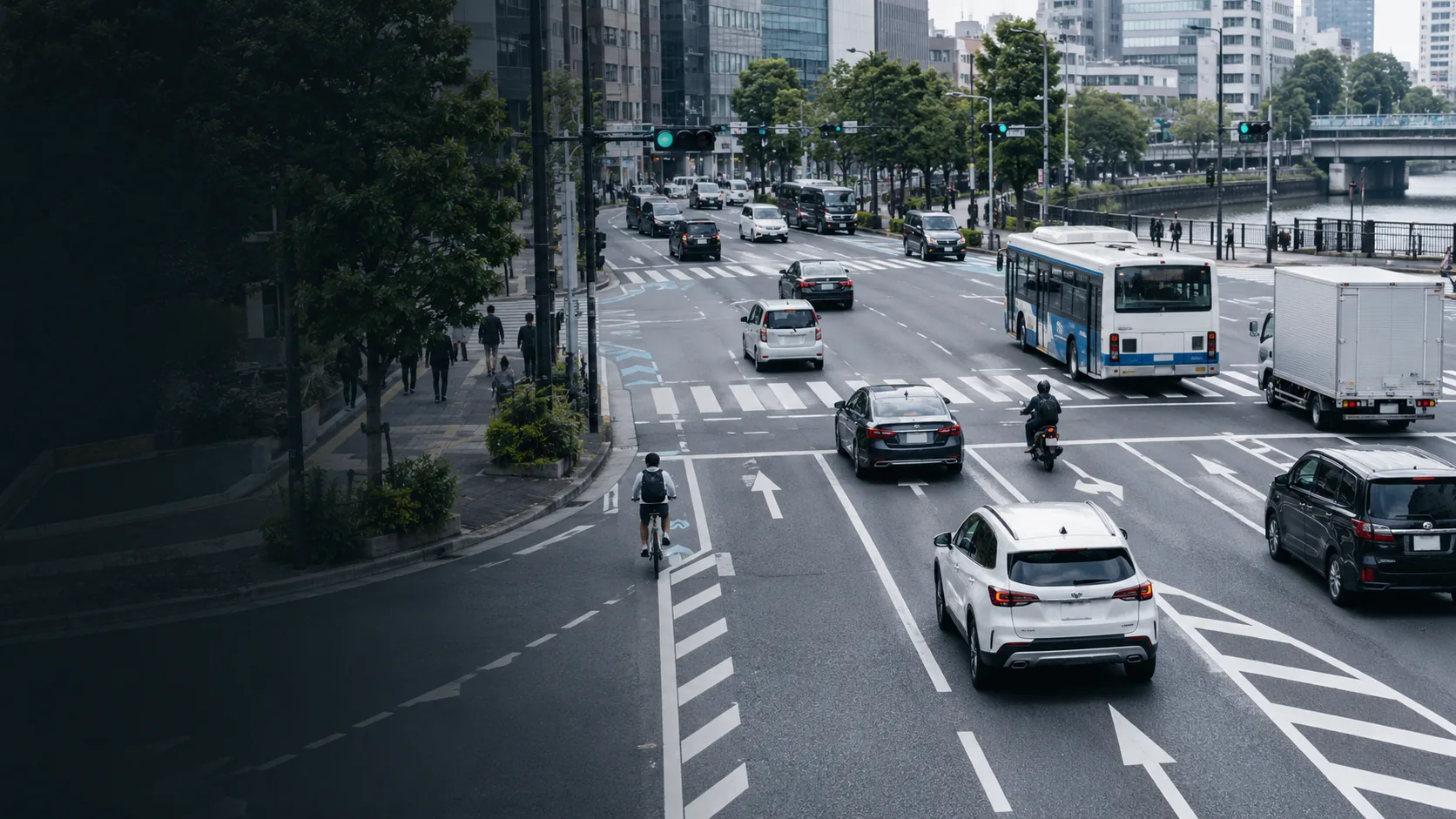 A multi-lane city street carrying cars, a bus, a truck, a motorcycle and a cyclist across marked lanes and a crossing